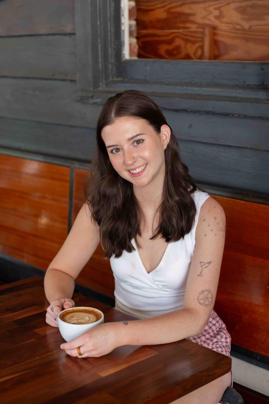 woman seated at a wooden table with a mug in her hands smiling.