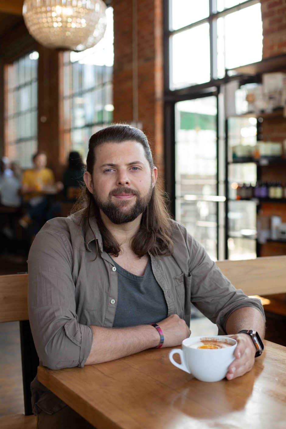 man with long hair and a beard seated at a table in a cafe with a mug in his hand.